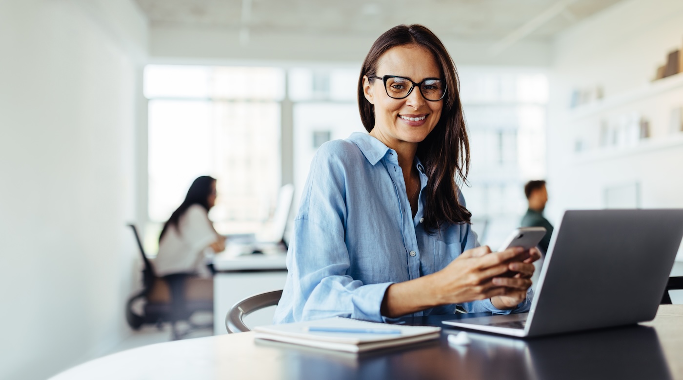 Mature business woman using a mobile phone while sitting in an office with a laptop. Woman working in a digital marketing agency.